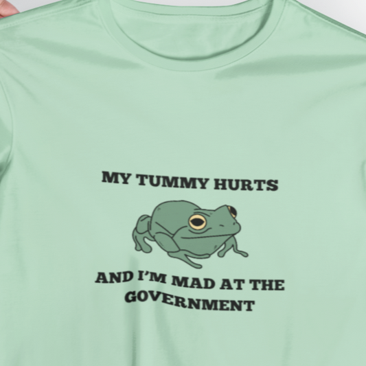 A green crew neck t-shirt with the text 'MY TUMMY HURTS AND I'M MAD AT THE GOVERNMENT' and an image of a frog in the center.