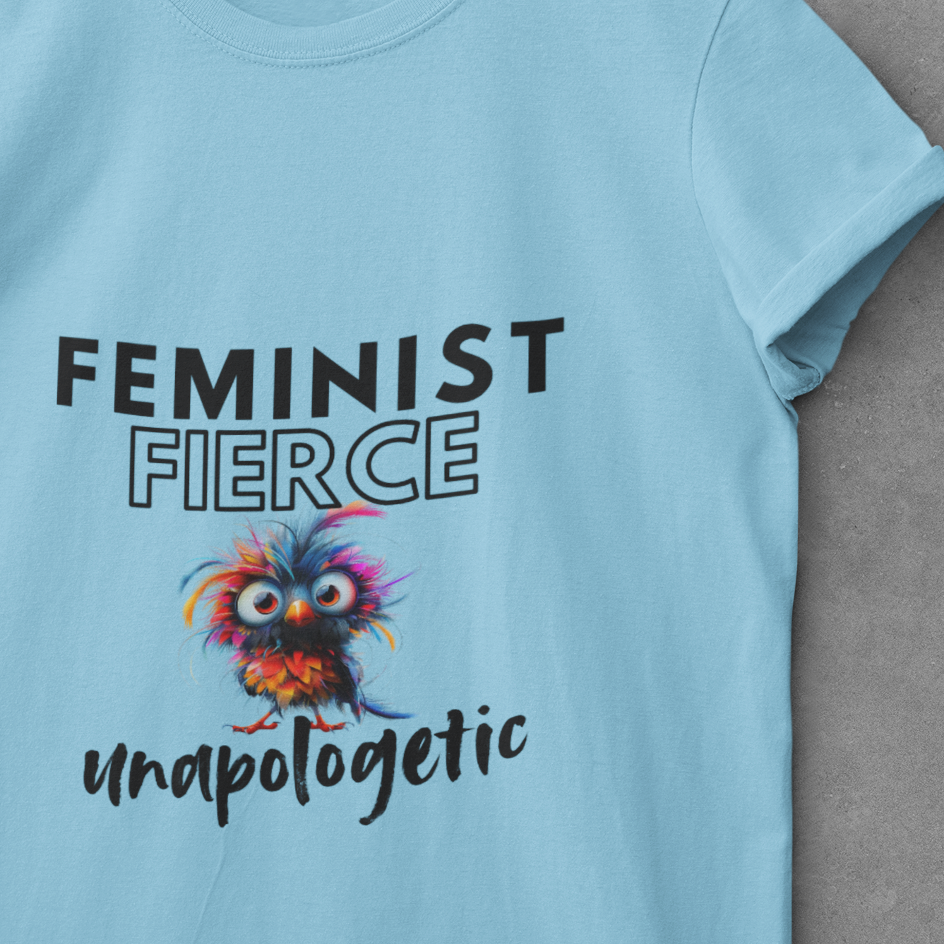 Light blue t-shirt with 'Feminist Fierce unapologetic' text and colorful owl graphic on a hanger against a gray background.
