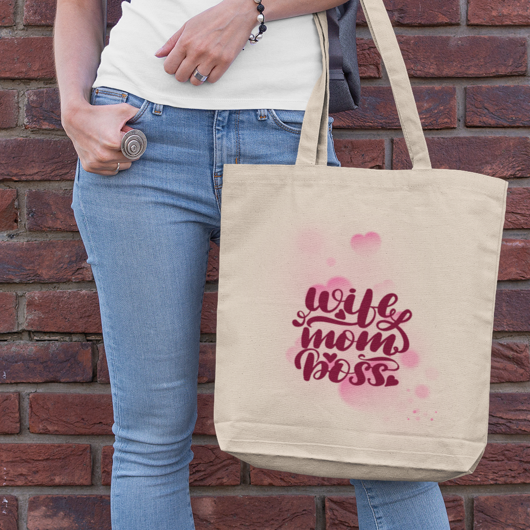 A person holding a beige tote bag with text on it, standing against a stone wall background.